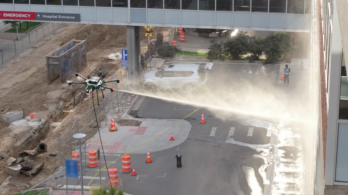 Seen from above, our cleaning drone sprays a hospital facade beside a construction zone; orange cones, caution tape and an observer in a safety vest mark the ground below.