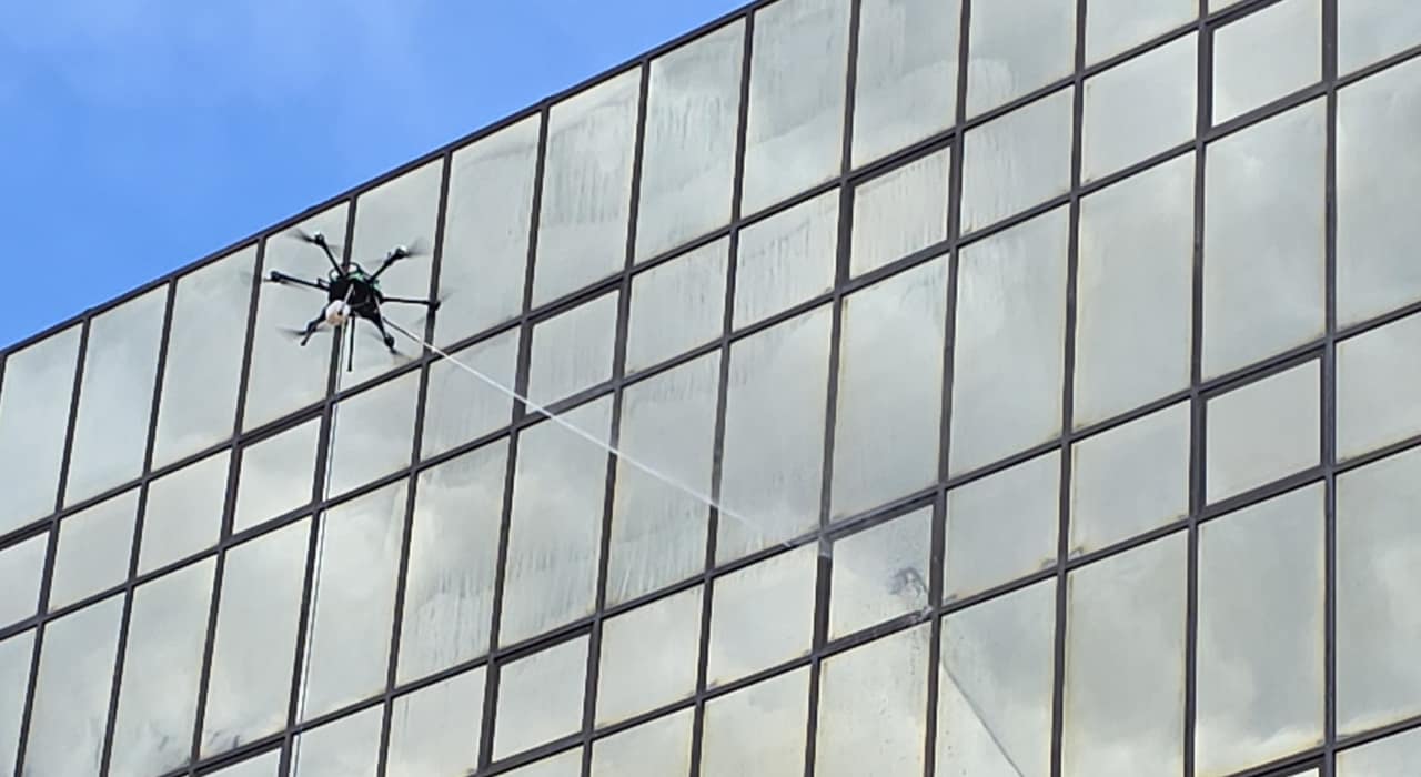 A cleaning drone directs a long jet across visibly hazed and spotted glass beneath a blue sky.