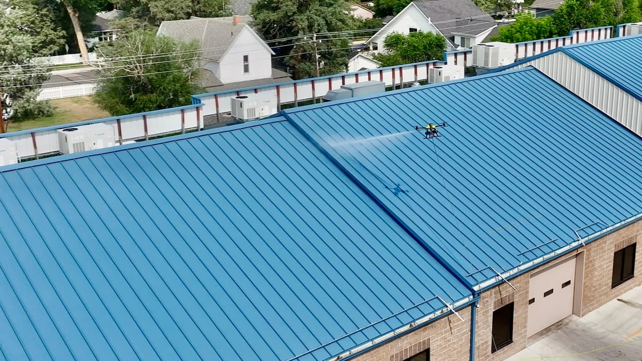 A tethered cleaning drone sprays a blue standing-seam metal roof, with its shadow visible on the panels below.