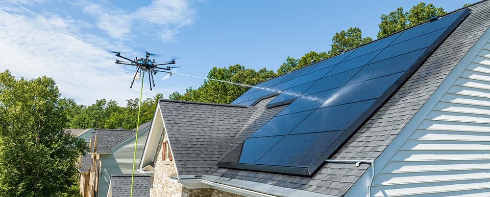 A tethered cleaning drone sprays water across solar panels on a sloped residential roof, with trees and blue sky behind it.