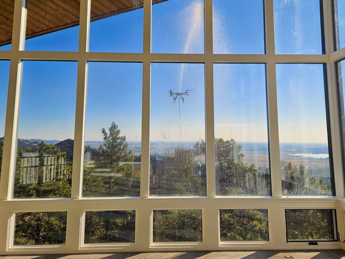 Seen from inside through tall white-framed windows, a drone outside sprays the glass against a blue sky.